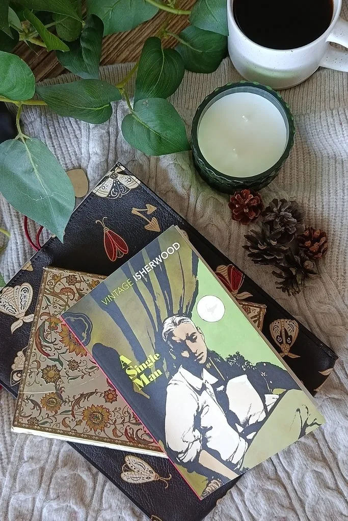 A photo of the paperback edition of A Single Man by Christopher Isherwood set against some journals, warm blankets and pinecones