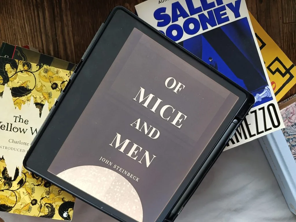A photo of Mice and Men by John Steinbeck open on Kindle Scribe, against a background of stacked books
