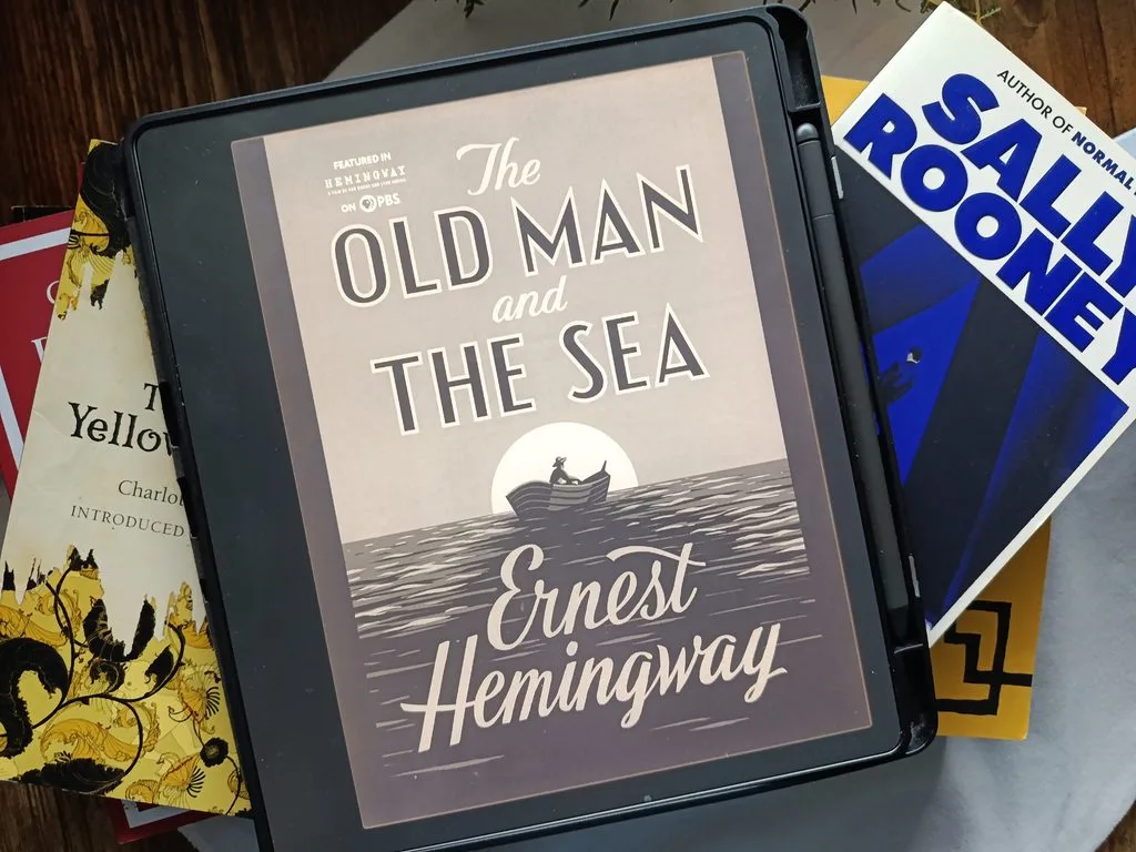 A photo of The Old Man and The Sea by Ernest Hemingway open on Kindle Scribe, propped up against stacked books