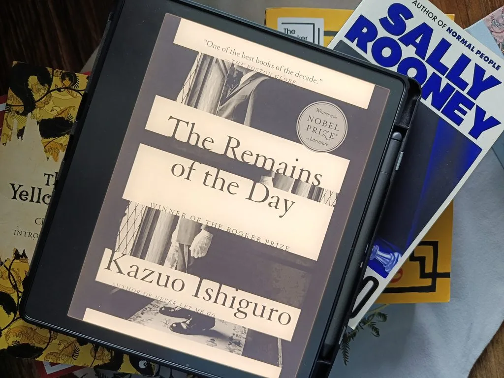 A photo of The Remains of The Day by Kazuo Ishiguro open on Kindle Scribe against a background of stacked books