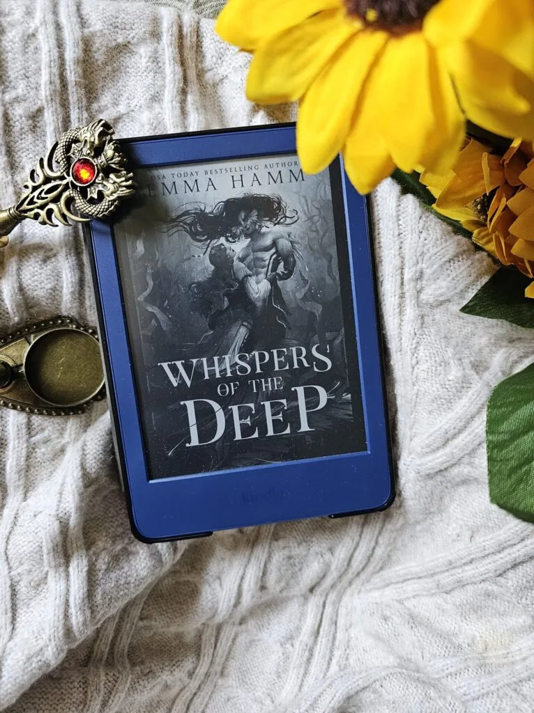 A photo of Whispers of The Deep by Emma Hamm open on a Kindle device so it's showing the cover, with some flowers and calligraphy items decorating the area around it
