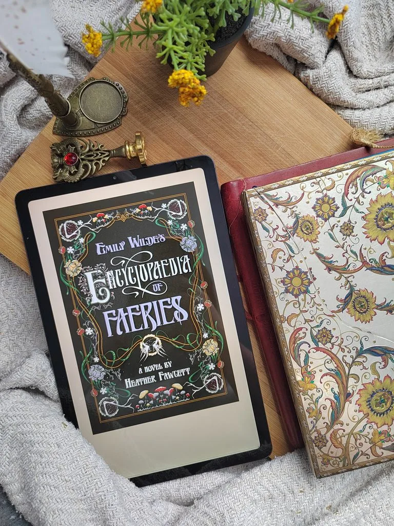 A photo of Emily Wilde's Encyclopaedia of Faeries open on a tablet, against a wooden board, with some journals for decor