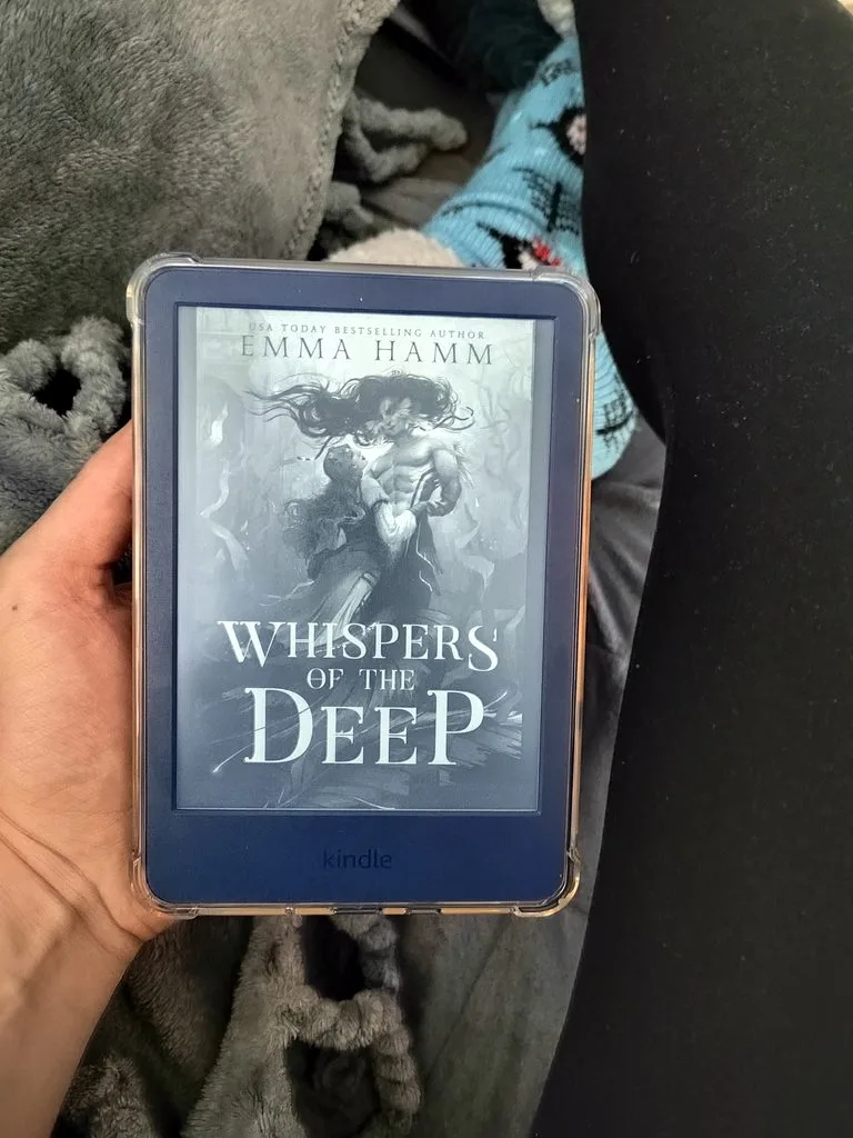 A photo of Whispers of The Deep open on Kindle, showing it held in a hand against a blanket