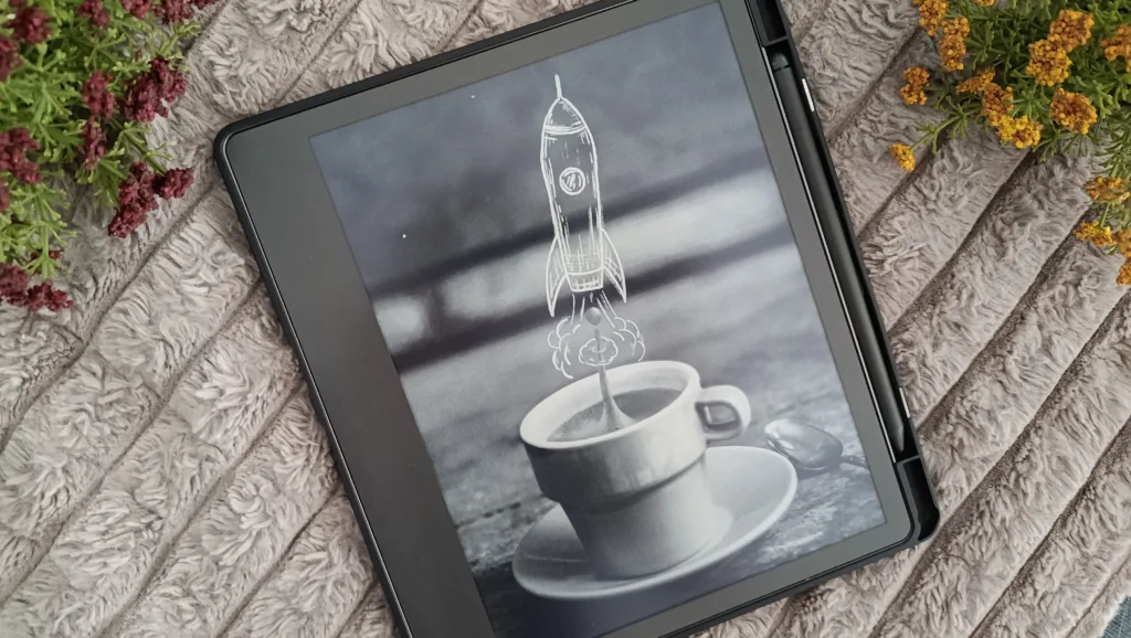 A photo of a Kindle Scribe locked, in a case and with the stylus at est, with flowers for decoration; the purpose of the photo is to illustrate what a kindle scribe looks like