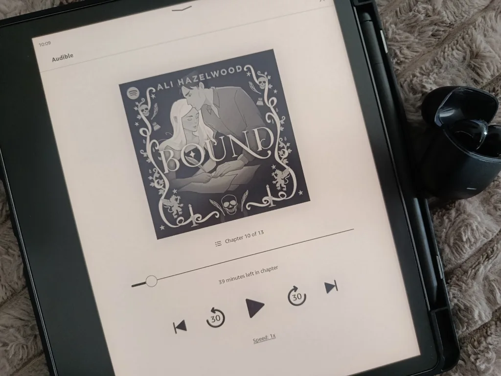 A photo of a kindle device showing an audiobook open and playing, with headphones next to it, showing how listening on Kindle works