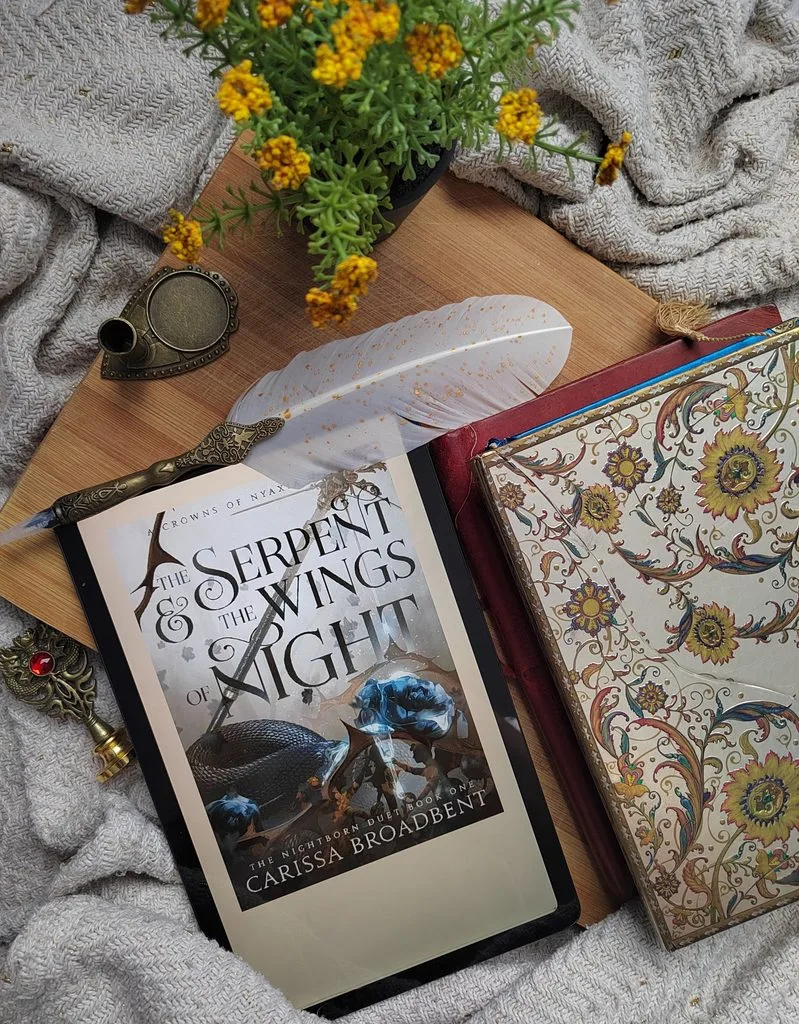 An e-book edition of The Serpent and the Wings of Night open on a tablet, with some journals, writing feather and cream blanket for decor