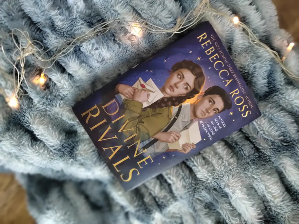 A photo of Divine Rivals by Rebecca Ross, the hardback edition with characters painted on, against a bluish gray blanket background and some string lights
